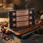 Three Satya Palo Santo blue boxes with smoking stick in wooden holder on dark stone with dried botanicals - Aroma Paradise